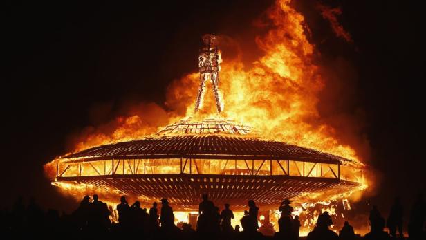 Das Burning Man Festival: Eine große Holzkonstruktion steht in Flammen, während Menschen im Dunkeln zusehen.