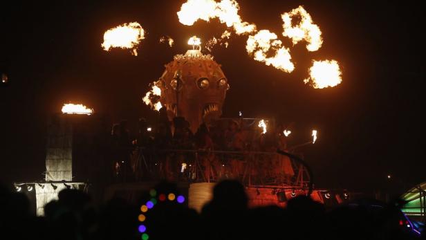 Eine große, feuerspeiende Skulptur auf einer Bühne bei Nacht zieht die Blicke auf sich.