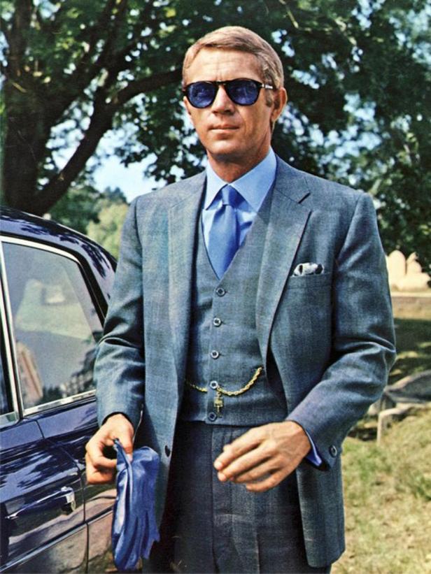 Steve McQueen in einem grauen Anzug mit blauen Akzenten und Sonnenbrille, neben einem Auto stehend.