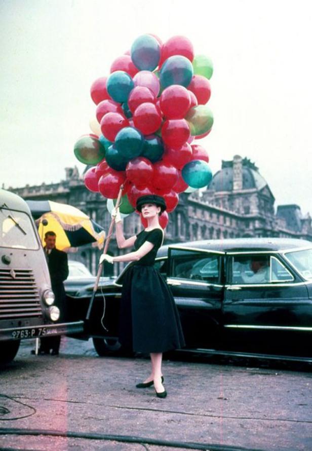 Eine Frau in einem schwarzen Kleid und Hut hält eine große Anzahl bunter Ballons.