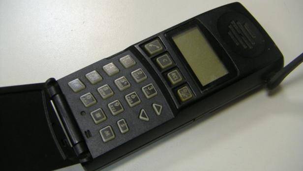 Ein älteres, schwarzes Klapphandy mit Antenne liegt auf einem Tisch.