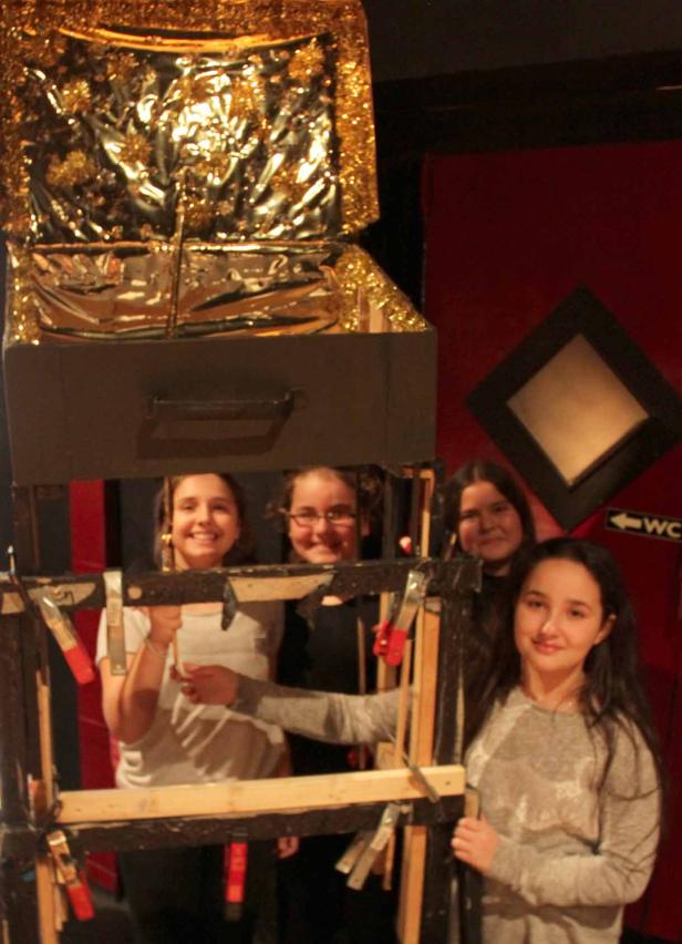 Vier junge Frauen stehen hinter einer hölzernen Rahmenkonstruktion mit einer glitzernden Kiste darüber.