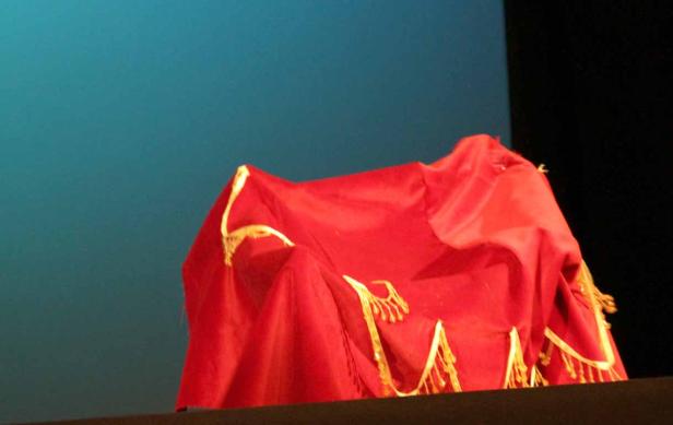 Ein rotes Tuch mit goldenen Verzierungen bedeckt etwas auf einer Bühne.