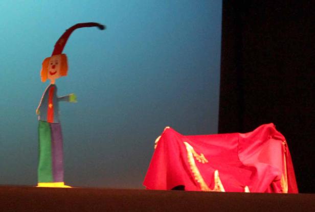 Eine Marionette in Clownskleidung steht neben einem roten Vorhang auf einer Bühne.