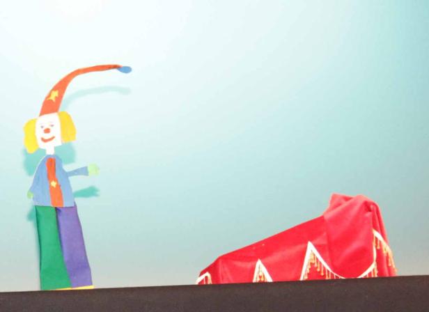 Eine Papierpuppe eines Clowns steht neben einem kleinen roten Zirkuszelt.