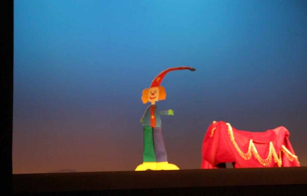 Eine Marionette mit Clown-Gesicht steht neben einem roten, drapierten Tisch.