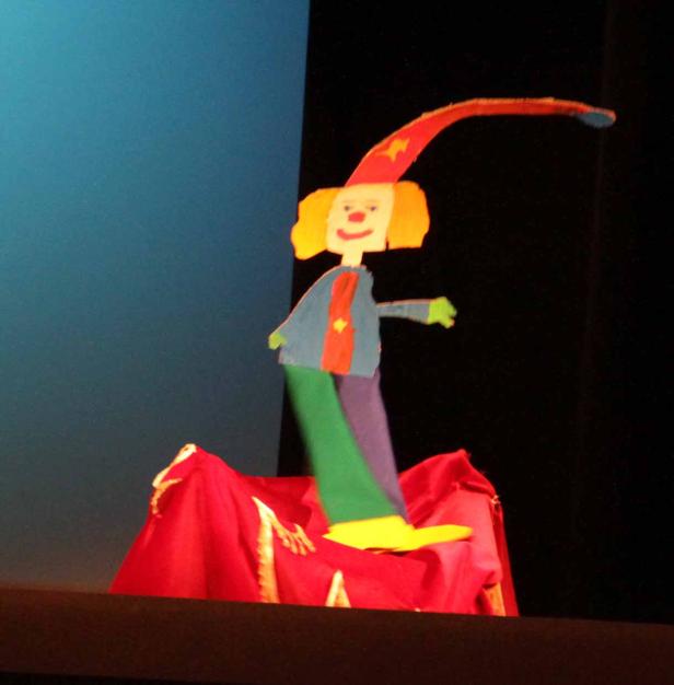 Eine bunte, selbstgemachte Clown-Figur steht auf einer roten Bühne.