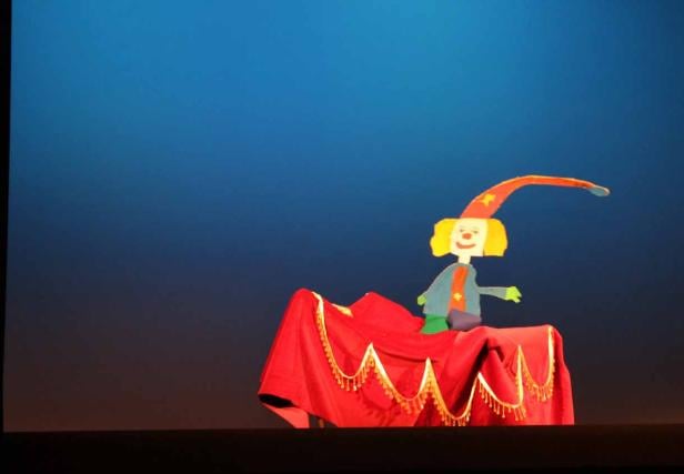 Eine Marionette in Form eines Clowns auf einer Bühne mit rotem Vorhang.