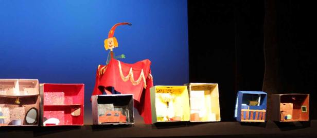 Eine Marionette steht auf einer Bühne vor einer Reihe von Miniatur-Szenen in Schuhkartons.