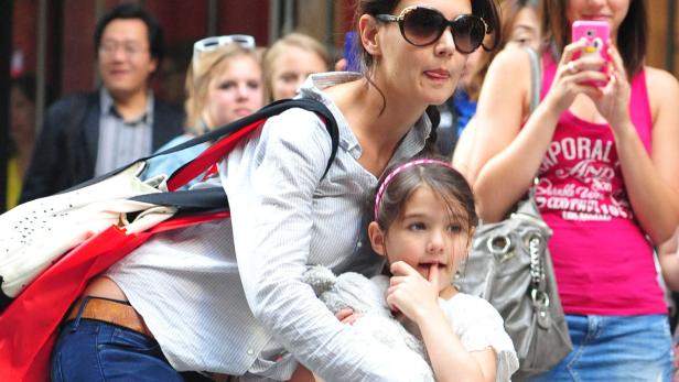 Katie Holmes trägt ihre Tochter Suri Cruise auf dem Arm, umgeben von Menschen.