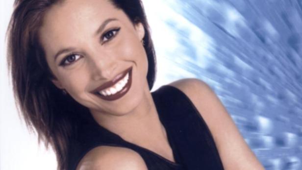 Eine lächelnde Frau mit dunklem Haar und dunklem Lippenstift vor einem blauen Hintergrund.