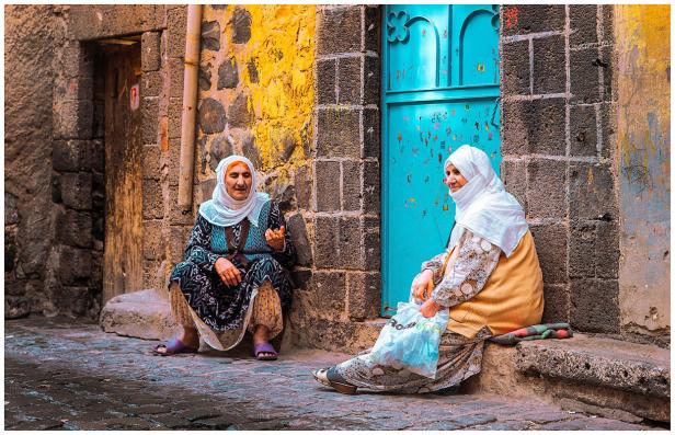Zwei ältere Frauen sitzen vor farbenfrohen Mauern in einer Gasse.