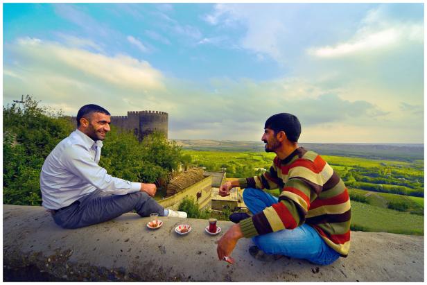 Zwei Männer sitzen auf einer Mauer mit Blick auf eine Burg und eine grüne Landschaft.