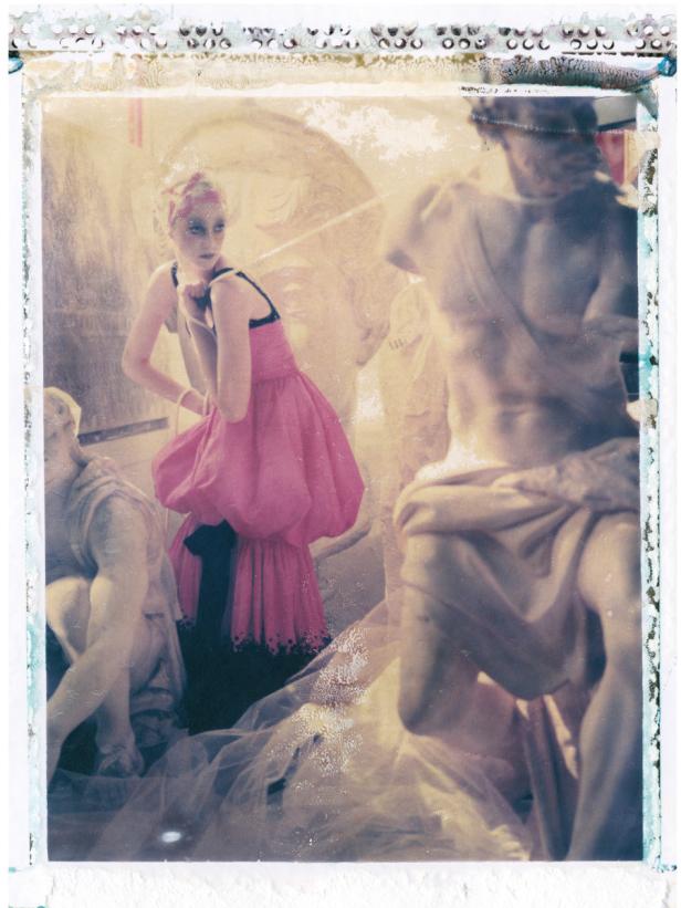 Eine Frau in einem rosa Kleid posiert zwischen antiken Statuen.