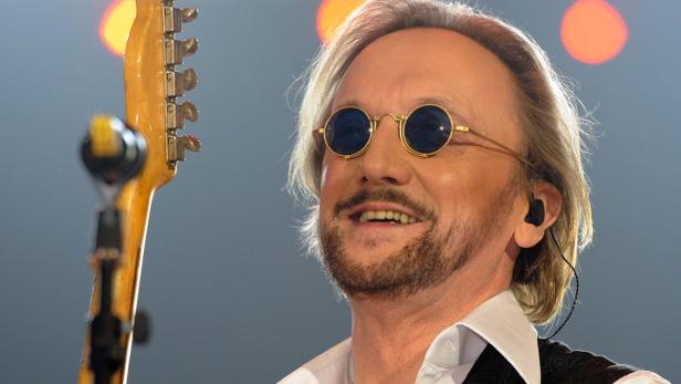 Ein Mann mit runden, blauen Brillengläsern lächelt neben dem Hals einer Gitarre.