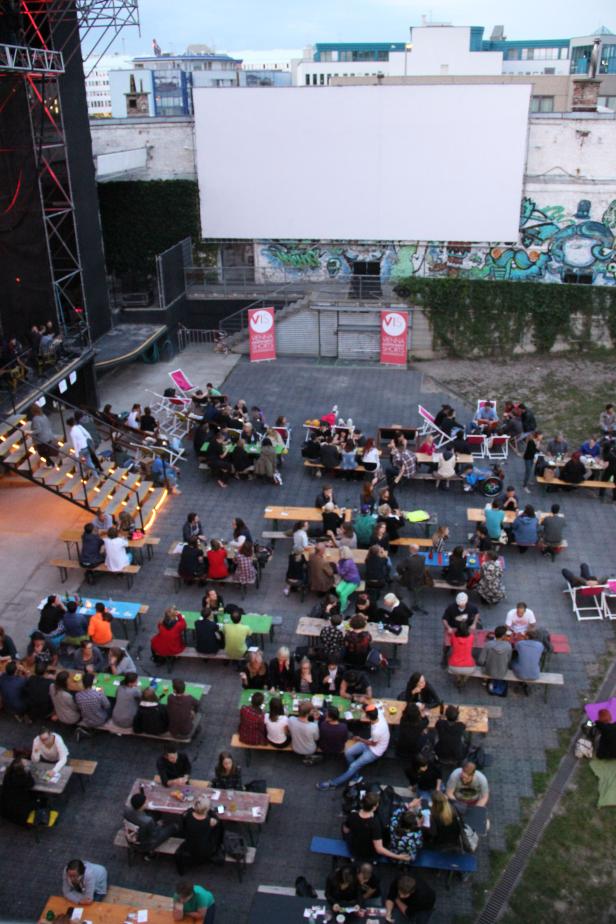 Ein Open-Air-Kino mit vielen Menschen an Tischen vor einer großen Leinwand.