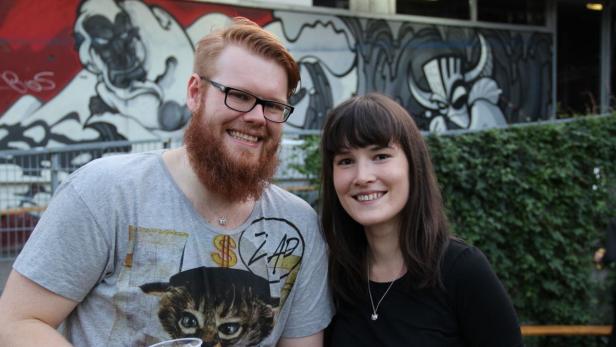 Ein Mann mit Bart und Brille und eine Frau lächeln vor einem Graffiti.