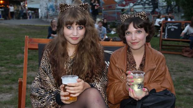 Zwei junge Frauen mit Leopardenmuster und Katzenohren sitzen mit Getränken im Freien.