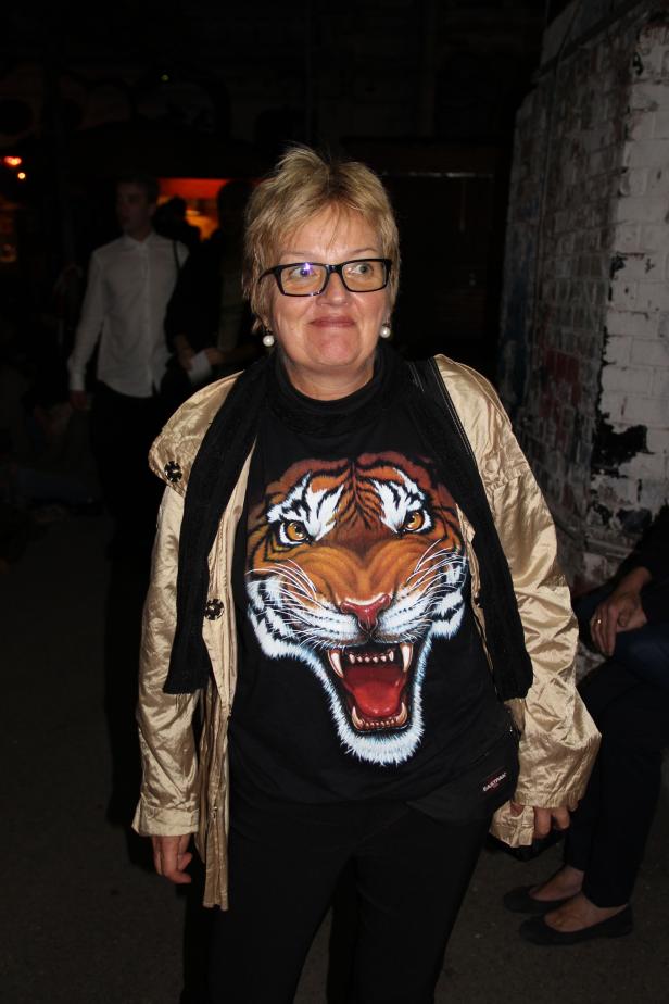 Eine Frau mit Brille und einem T-Shirt mit Tigerdruck steht vor einer Wand.