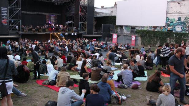Viele Menschen sitzen oder stehen bei einer Open-Air-Veranstaltung vor einer Bühne.