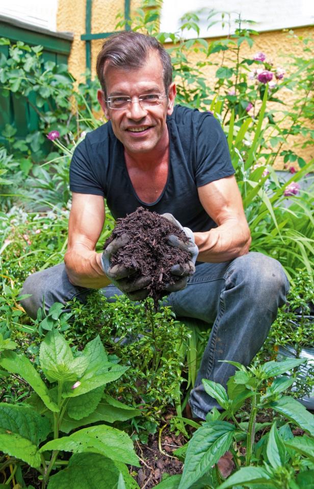 Ein Mann hockt im Garten und hält eine Handvoll Erde in die Kamera.