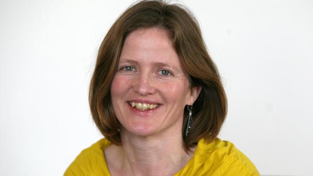 Eine lächelnde Frau mit braunen Haaren und einem gelben Oberteil vor einem weißen Hintergrund.