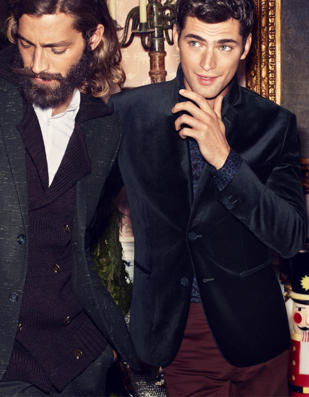 Zwei Männer posieren in eleganten Anzügen vor einem festlichen Hintergrund.