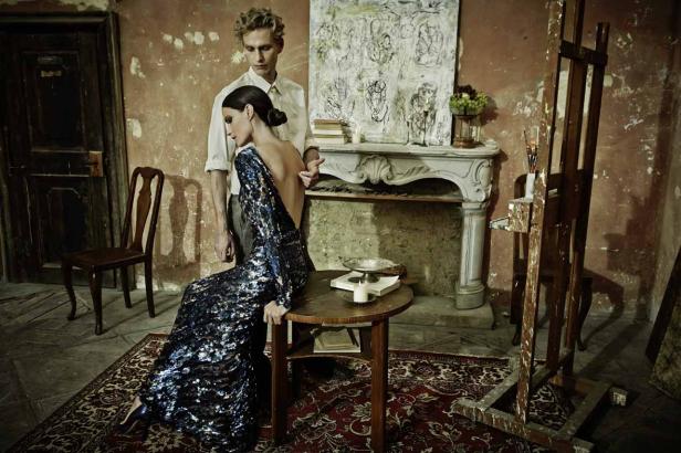 Eine Frau im Paillettenkleid posiert in einem Atelier, während ein Mann hinter ihr steht.