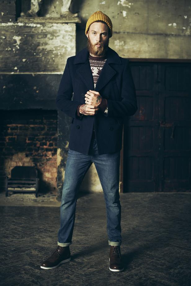 Ein bärtiger Mann mit Mütze, Jeans und Cabanmantel steht vor einer Backsteinmauer.