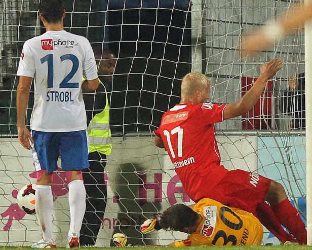 Ein Fußballspiel: Ein Spieler in Rot erzielt ein Tor, während der Torwart am Boden liegt.