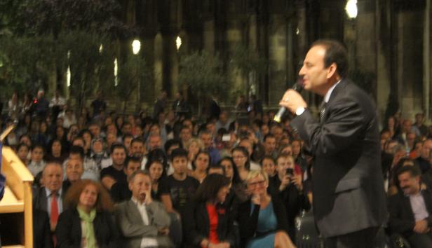 Ein Mann in Anzug spricht vor einer großen Menschenmenge in einer Außenanlage.