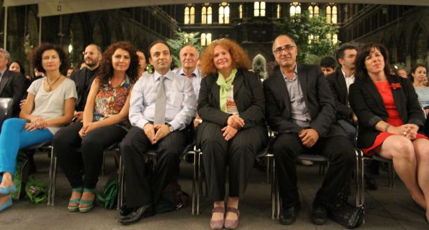 Eine Gruppe von Menschen sitzt bei einer Veranstaltung vor einem Gebäude mit vielen Fenstern.