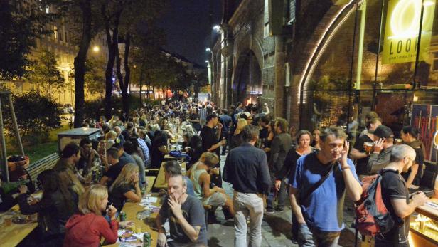 Viele Menschen sitzen und stehen an Tischen vor einem Lokal am Abend.