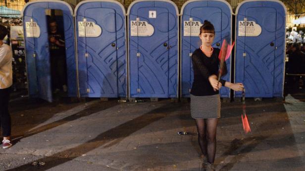 Eine Frau jongliert mit leuchtenden Stäben vor einer Reihe blauer Toilettenkabinen.