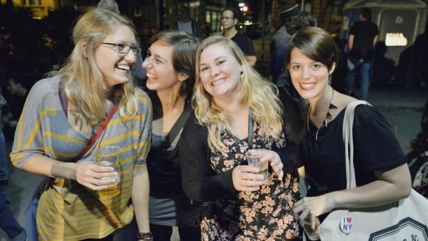 Vier junge Frauen stehen draußen und lachen, jede hält ein Getränk in der Hand.
