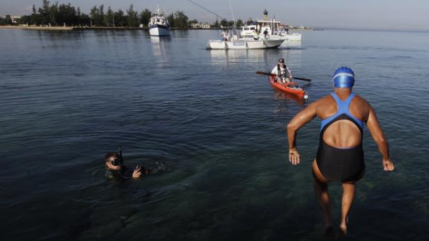 Eine Frau springt ins Wasser, während ein Taucher und mehrere Boote im Hintergrund zu sehen sind.