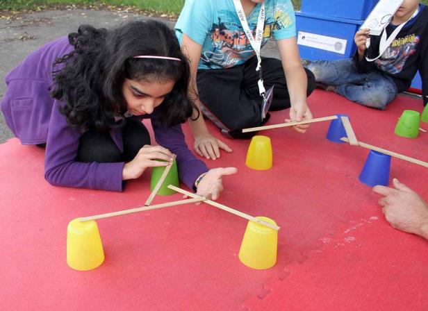 Kinder balancieren Holzstäbe auf Plastikbechern.