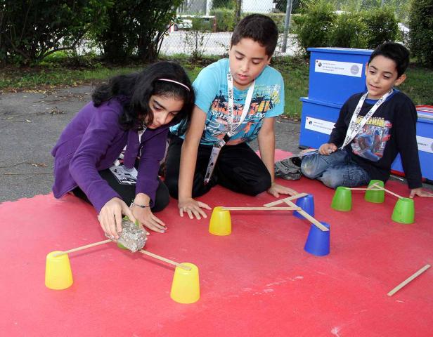 Drei Kinder bauen eine Brücke mit Bechern und Holzstäben.