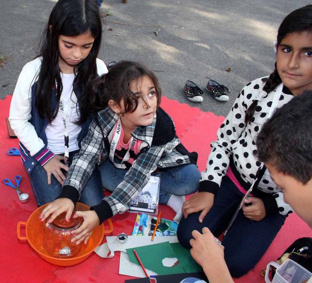 Eine Gruppe Kinder sitzt auf einer roten Matte und bastelt mit Papier und einer orangefarbenen Schüssel.