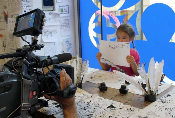 Ein Mädchen hält ein Blatt Papier mit der Aufschrift „Asht“. Eine Kamera filmt die Szene.