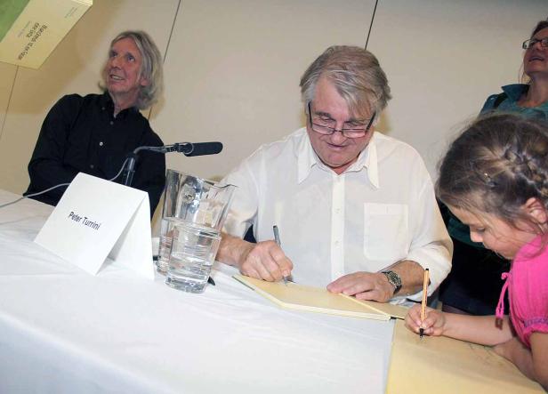 Peter Turrini signiert ein Buch, während ein junges Mädchen daneben auch unterschreibt.