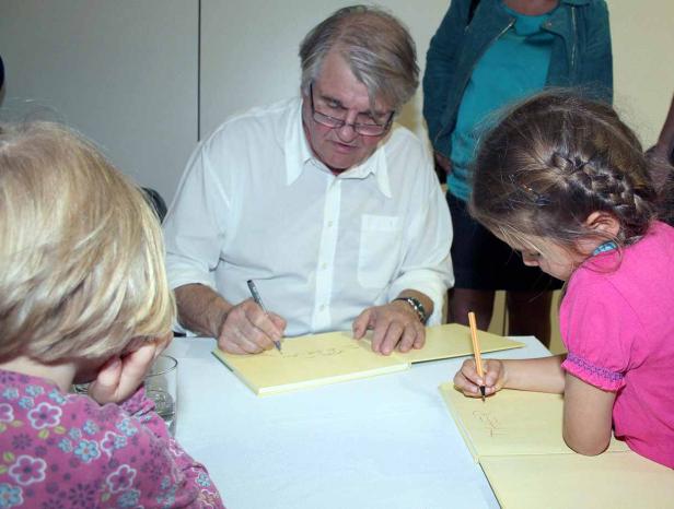 Ein Mann mit Brille signiert Bücher, während ein kleines Mädchen daneben malt.