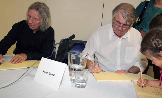 Peter Turrini signiert Bücher, während ein Mädchen daneben ebenfalls schreibt.