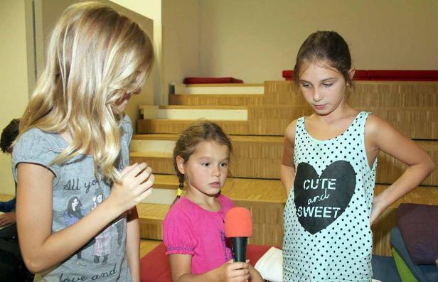 Drei Mädchen stehen auf einer Bühne, eines hält ein Mikrofon.