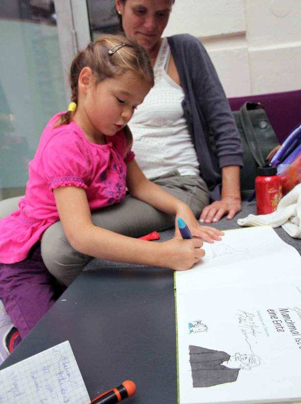 Ein junges Mädchen zeichnet mit einem blauen Stift in ein Buch, während eine Frau zusieht.