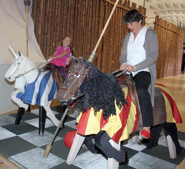 Eine Frau und ein Mädchen reiten auf hölzernen Steckenpferden mit Lanzen in einem Innenraum.