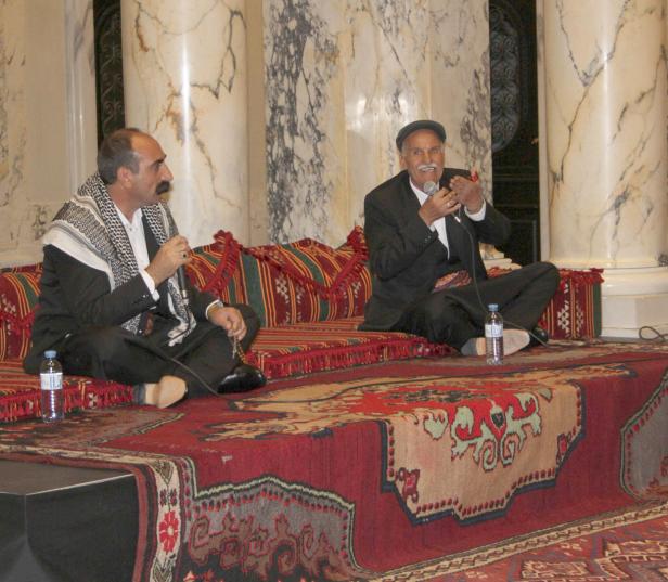 Zwei Männer sitzen auf einem verzierten Teppich und sprechen in Mikrofone.