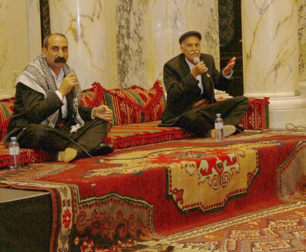 Zwei Männer sitzen auf einem verzierten Teppich und sprechen in Mikrofone.