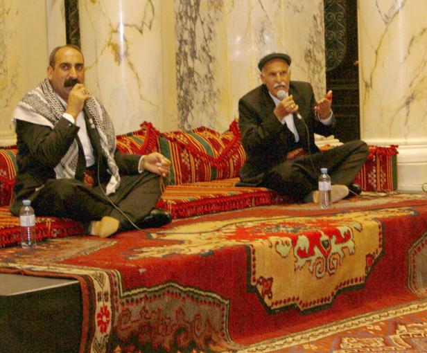 Zwei Männer sitzen auf einem verzierten Teppich und sprechen in Mikrofone.