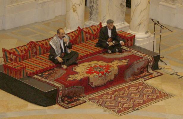 Zwei Männer sitzen auf einem traditionellen Sofa und sprechen in Mikrofone.